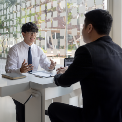 Two men sitting across a desk from one another during a job interview. One is wearing a white button-up shirt and blue tie, and the other is in a black jacket. An open laptop displaying a resume is on the desk, facing the man in the black jacket.