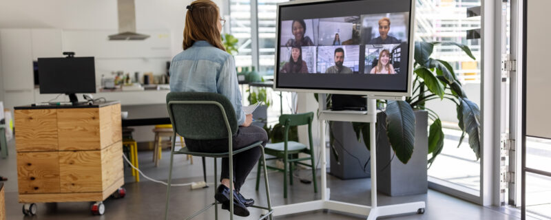 Woman sits in an office set up. In front of her, there is a meeting on her monitor with six people displayed.